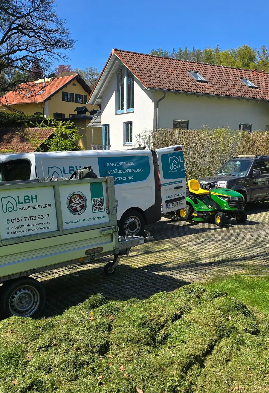 Einsatzwagen der LBH Hausmeisterei mit Anhänger und Aufsitzmäher vor einem Wohnhaus, davor frisch gemähtes Gras
